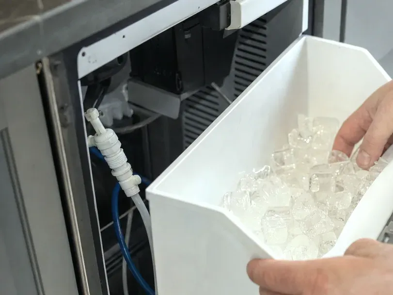 Hand holding a white ice bin full of cubes beside the ice maker's water line fitting and blue supply tubing.