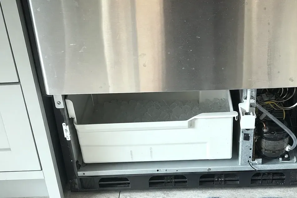 Stainless undercounter unit with its lower front removed, the white ice bin and refrigeration parts exposed.